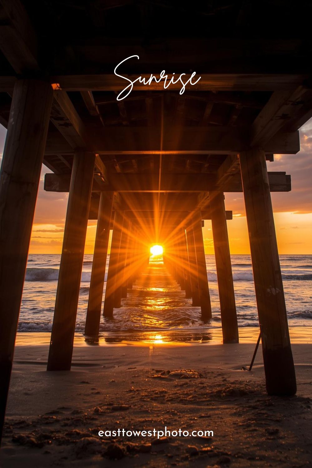 Sunrise under a beach pier with golden starburst rays reflecting on wet sand — canvas wall art by East to West Photo