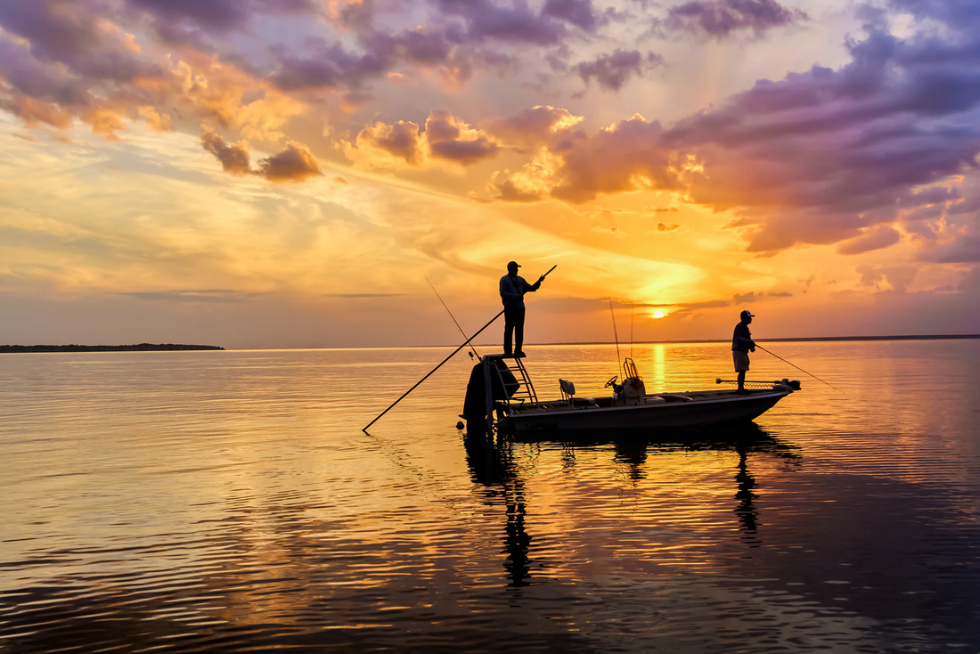 Two anglers silhouetted on a flat-bottomed skiff against a dramatic golden and purple sunset over calm coastal waters
